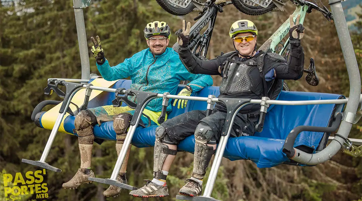 Two mountain bikers sitting on a chair lift, doing victory signs with their hands and grinning. Their bikes are hanging from the back of the chair lift.