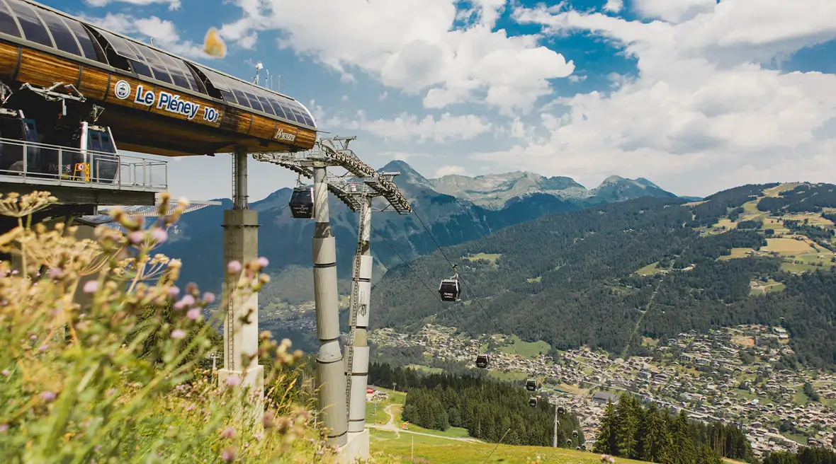 The view from a spot next to the top cable car station on Pleney, looking across Morzine in the far distance over to the green mountains beyond, against a sunny sky