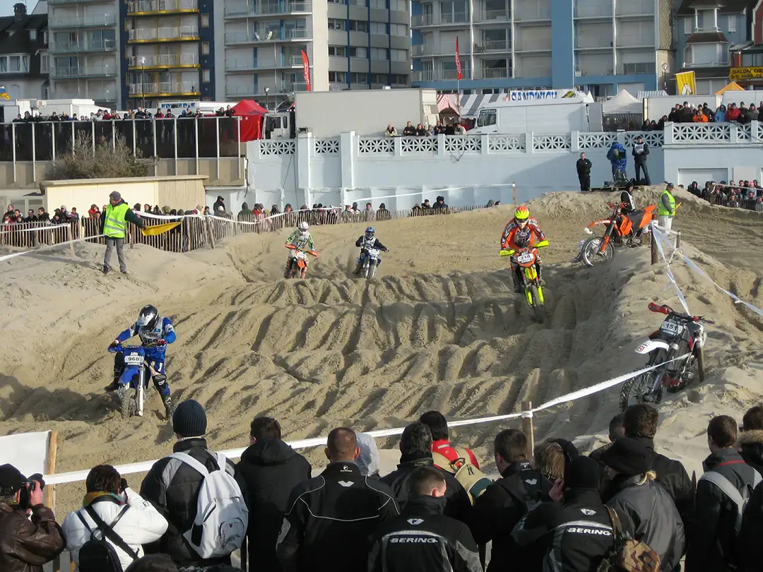 A group of motorbikes racing on sand mounds, with a few spectators watching and stewards.