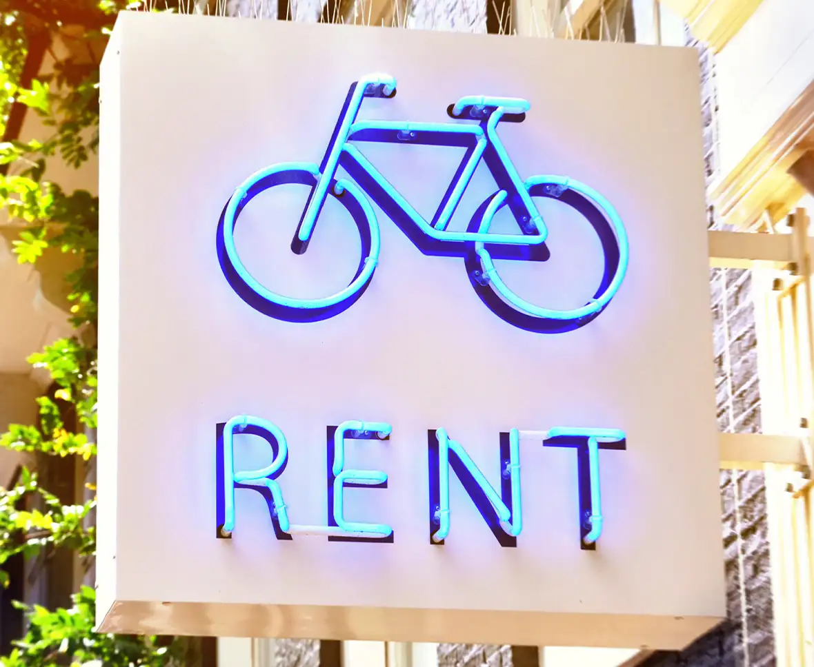 Blue lit up neon sign on a building that says Rent with a picture of a bike below