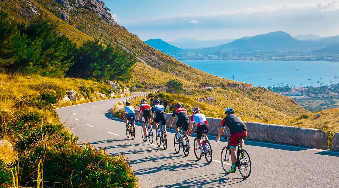 A group of amateur cyclists ride along a low level mountain road with an attractive sunlit bay in the background and a mountain range in the distance
