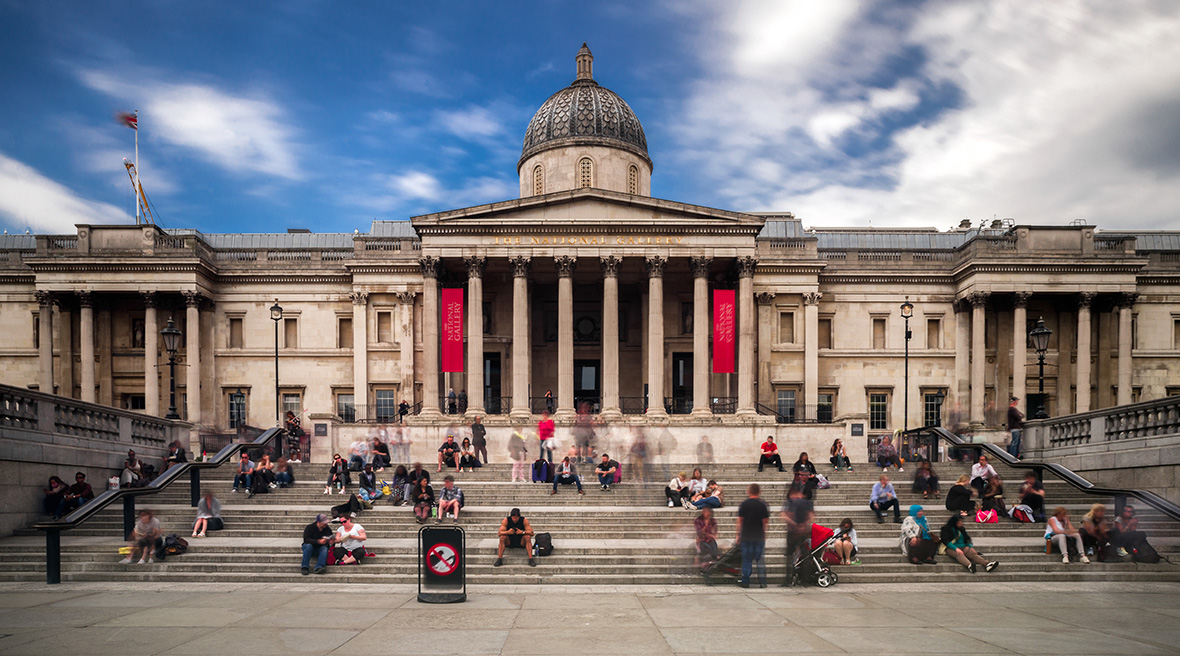 Die National Gallery mit hohen Säulen, roten Bannern und Menschen, die unter strahlend blauem Himmel auf den Treppen sitzen.
