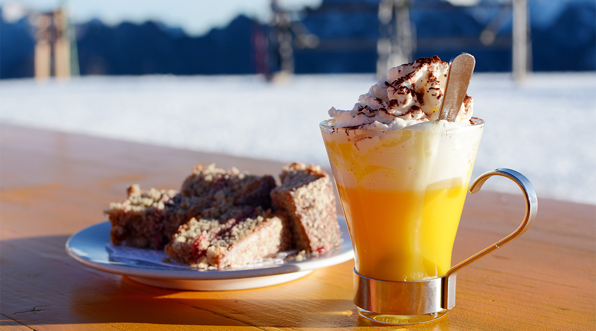 An egg coloured drink with a creamy head and a plate of cake on an outside bar table