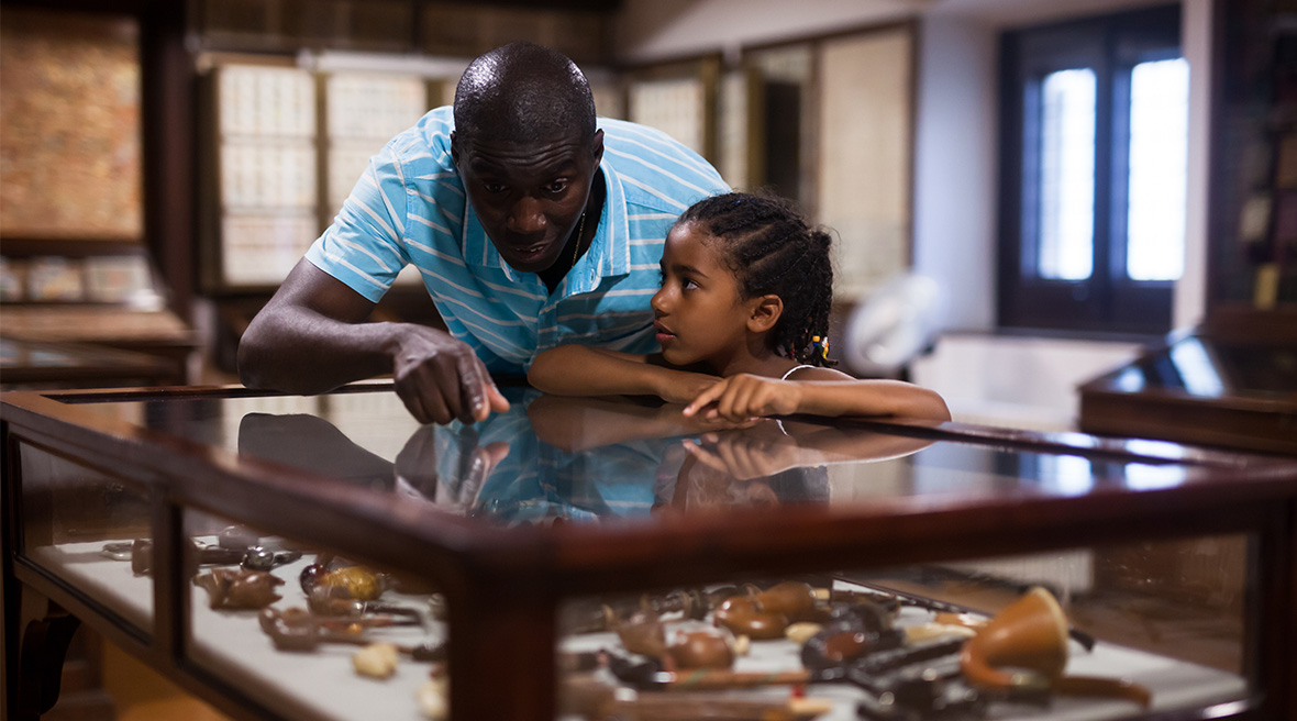 Ein Vater und seine Tochter betrachten ein Ausstellungsstück in einer Glasvitrine, zeigen auf etwas und unterhalten sich darüber.