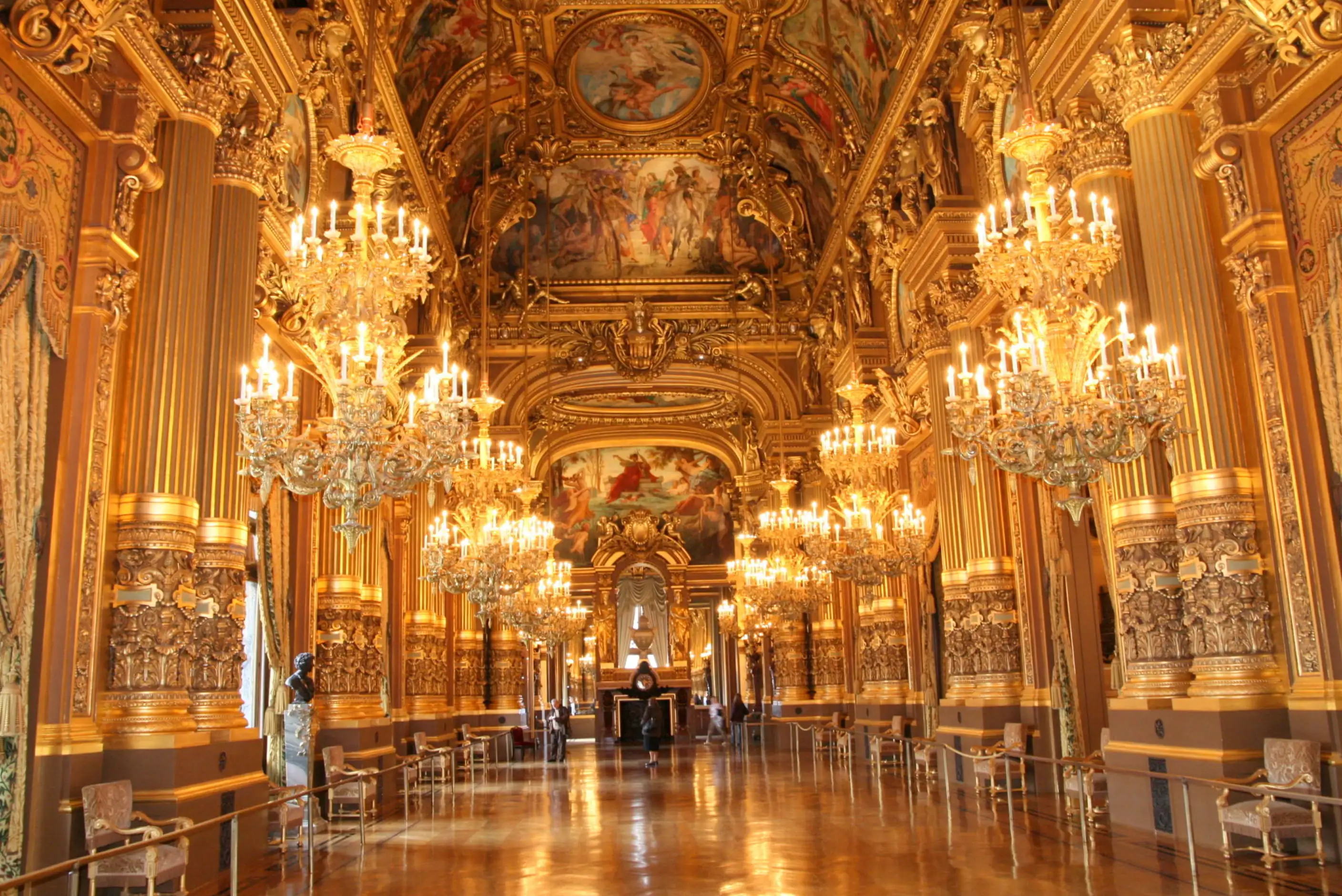 Interior of Palais Garnier, Paris, featuring gold decorations, crystal chandeliers, and ornate frescoes along a lavish hallway.