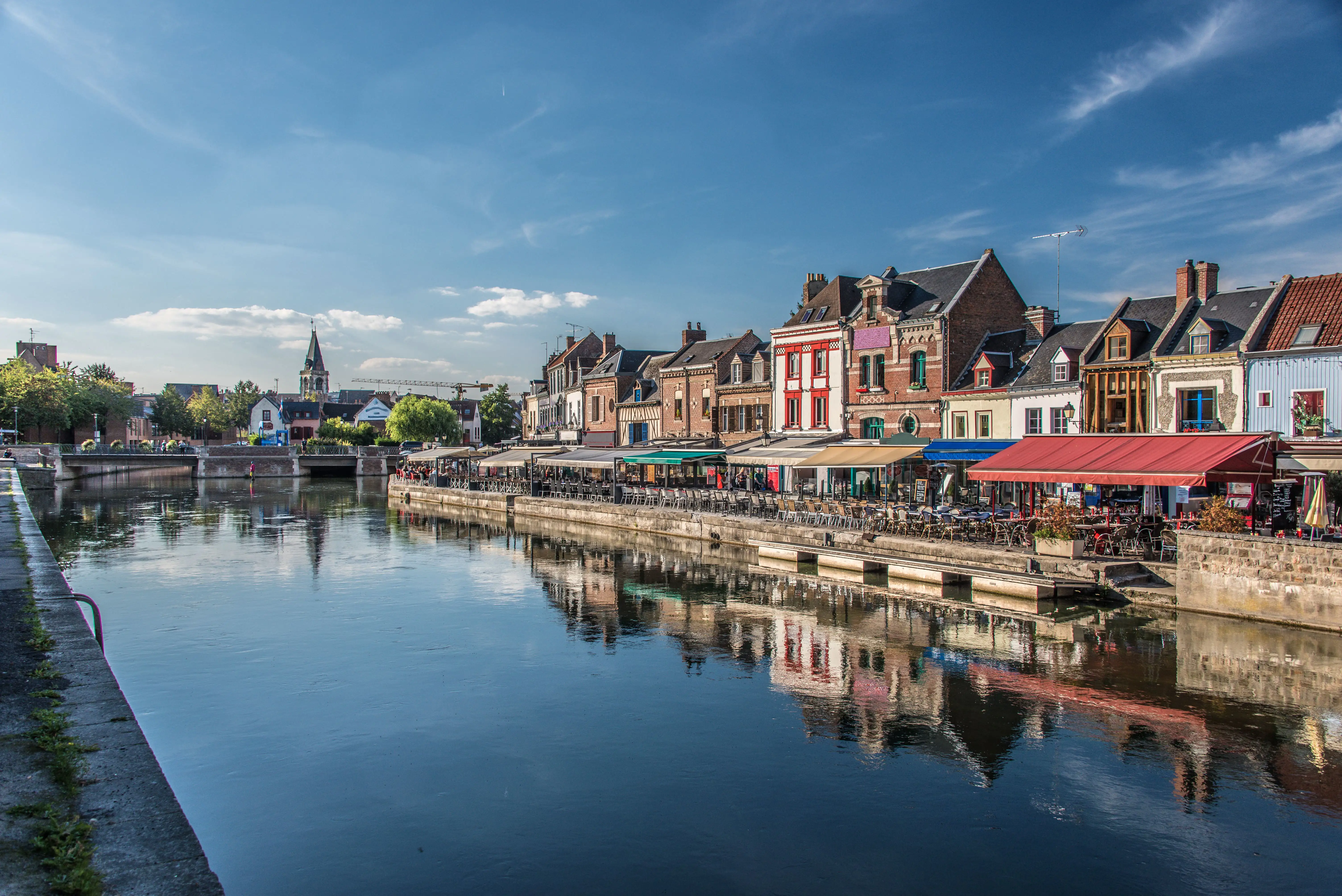 Quai Belu in Amiens with a canal, market stalls, and people under a bright blue sky.
