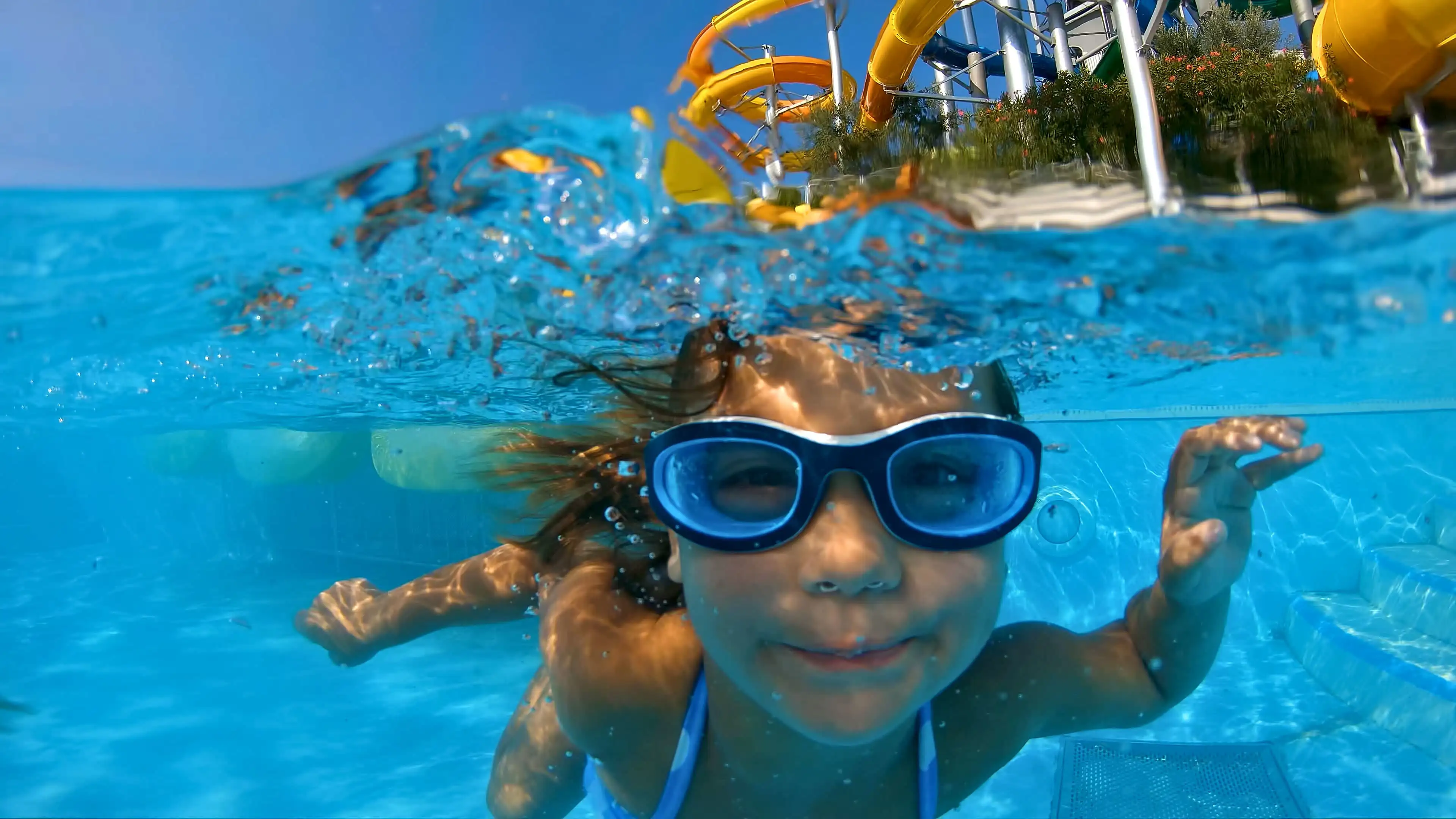A girl swimming underwater with goggles on and a slide in the background.