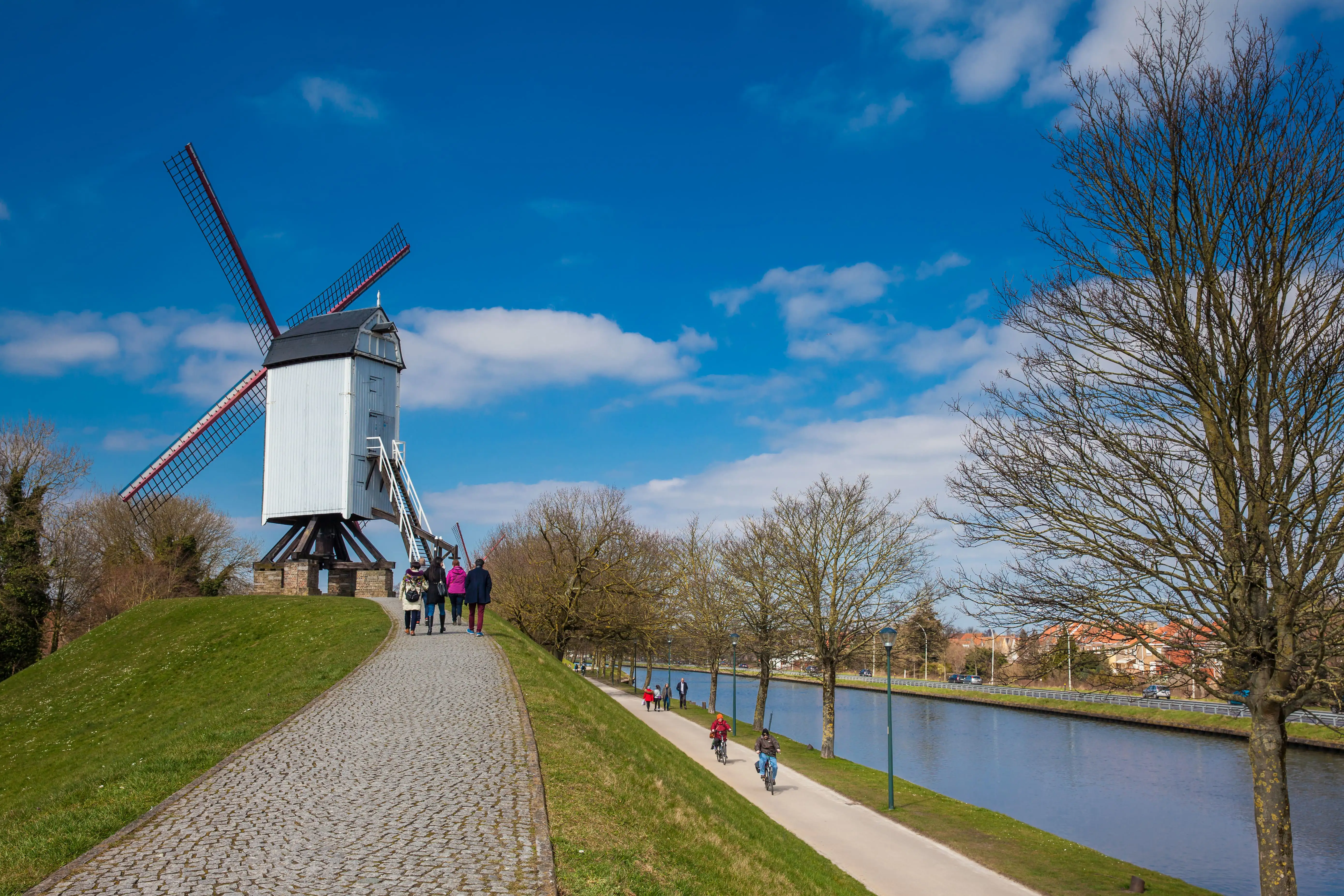 Paths, lined by grass, with a canal running along their side. A windmill is in the distance and walkers and cyclists pass by.