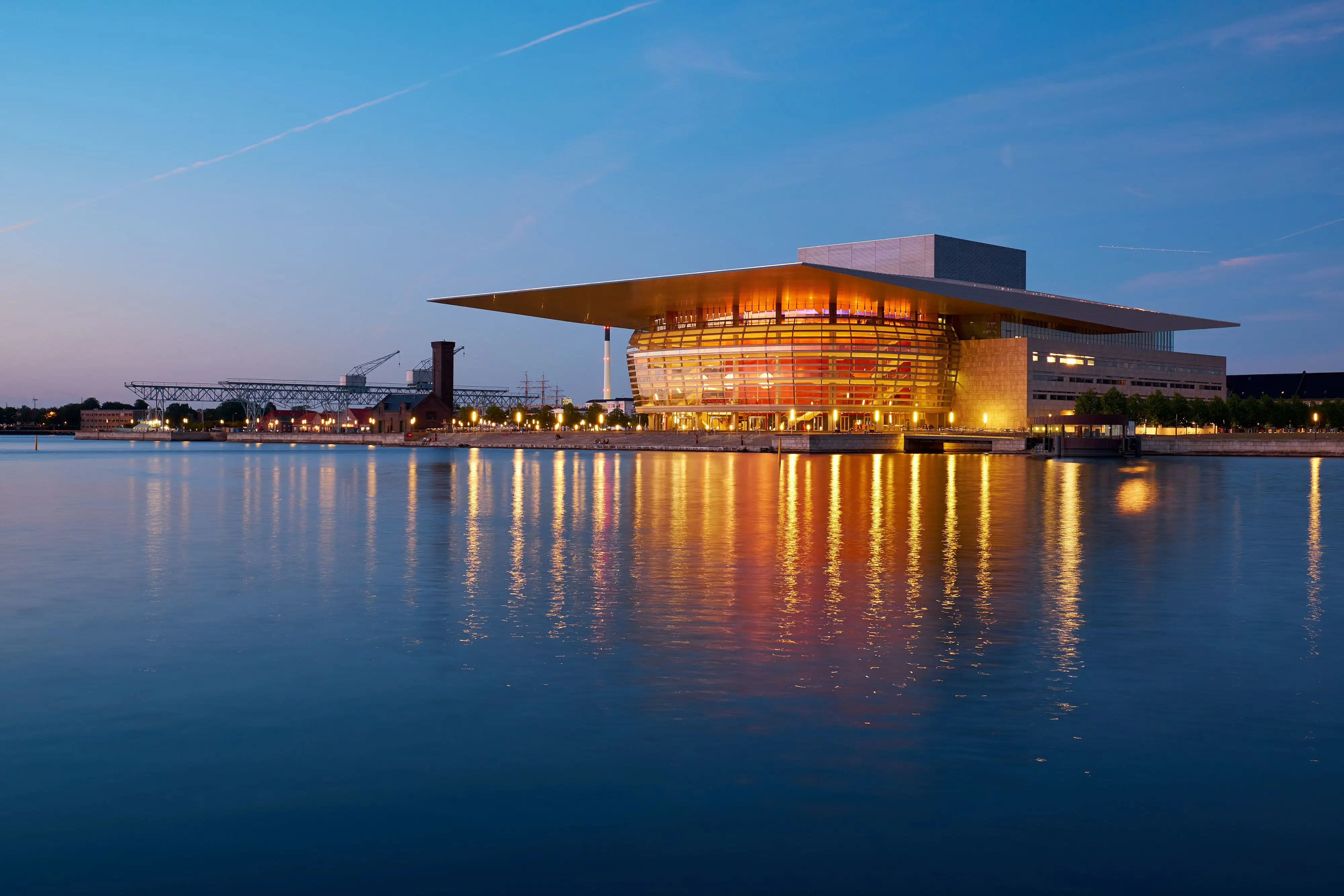 Copenhagen Opera House by the water at evening, glowing with warm lights.