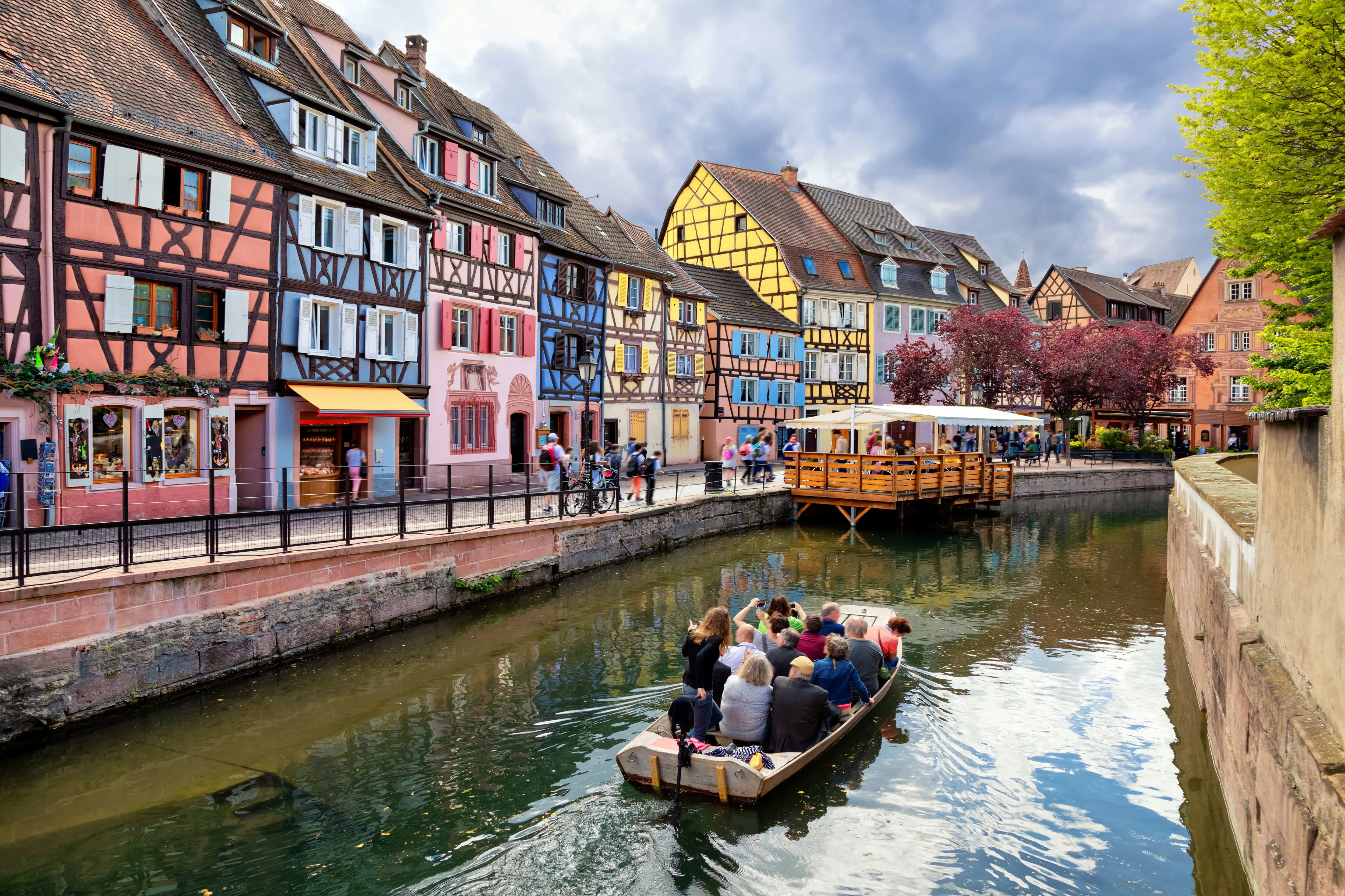 People enjoying a boat ride on the Lauch River in Colmar, with colourful half-timbered buildings lining the canal under a cloudy sky.