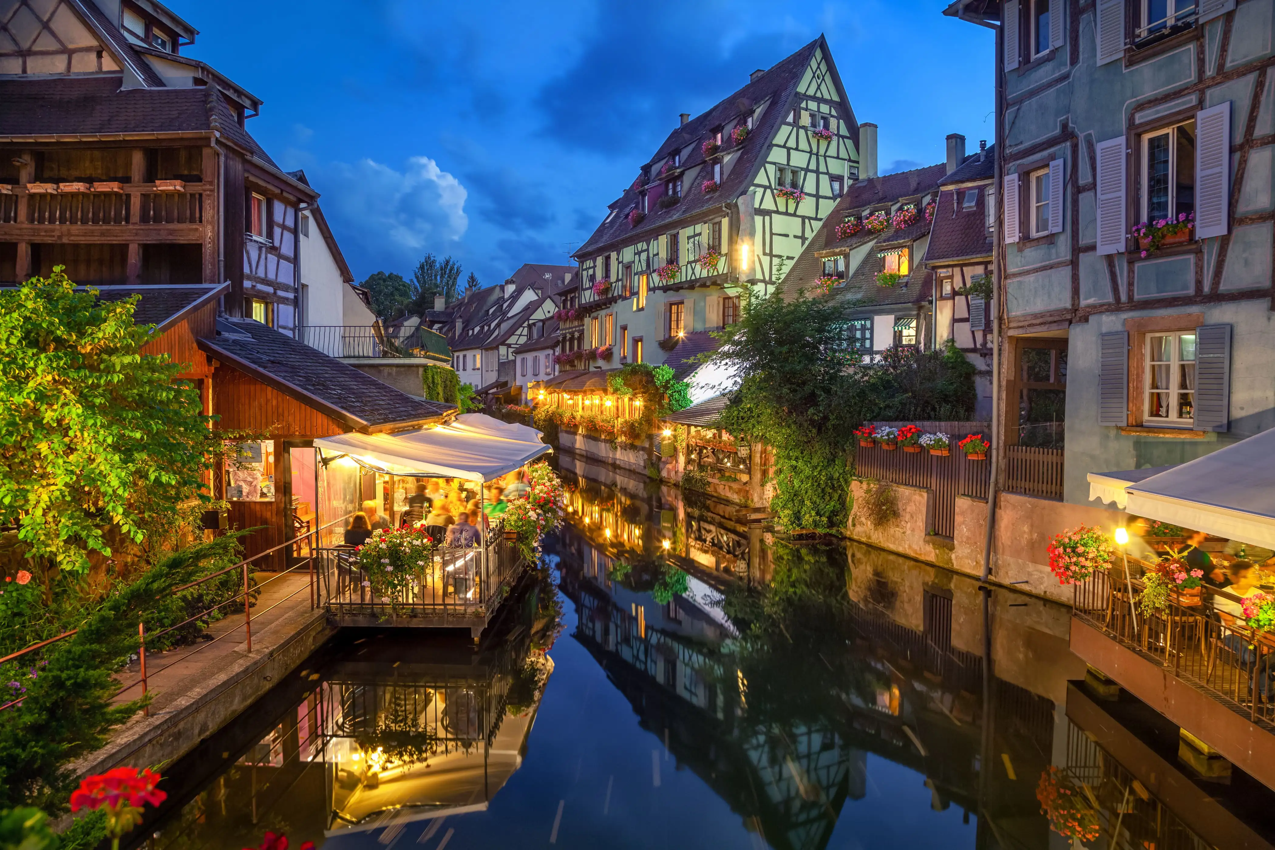Half-timbered houses and restaurant verandas with warm lights reflecting in the calm water at dusk.