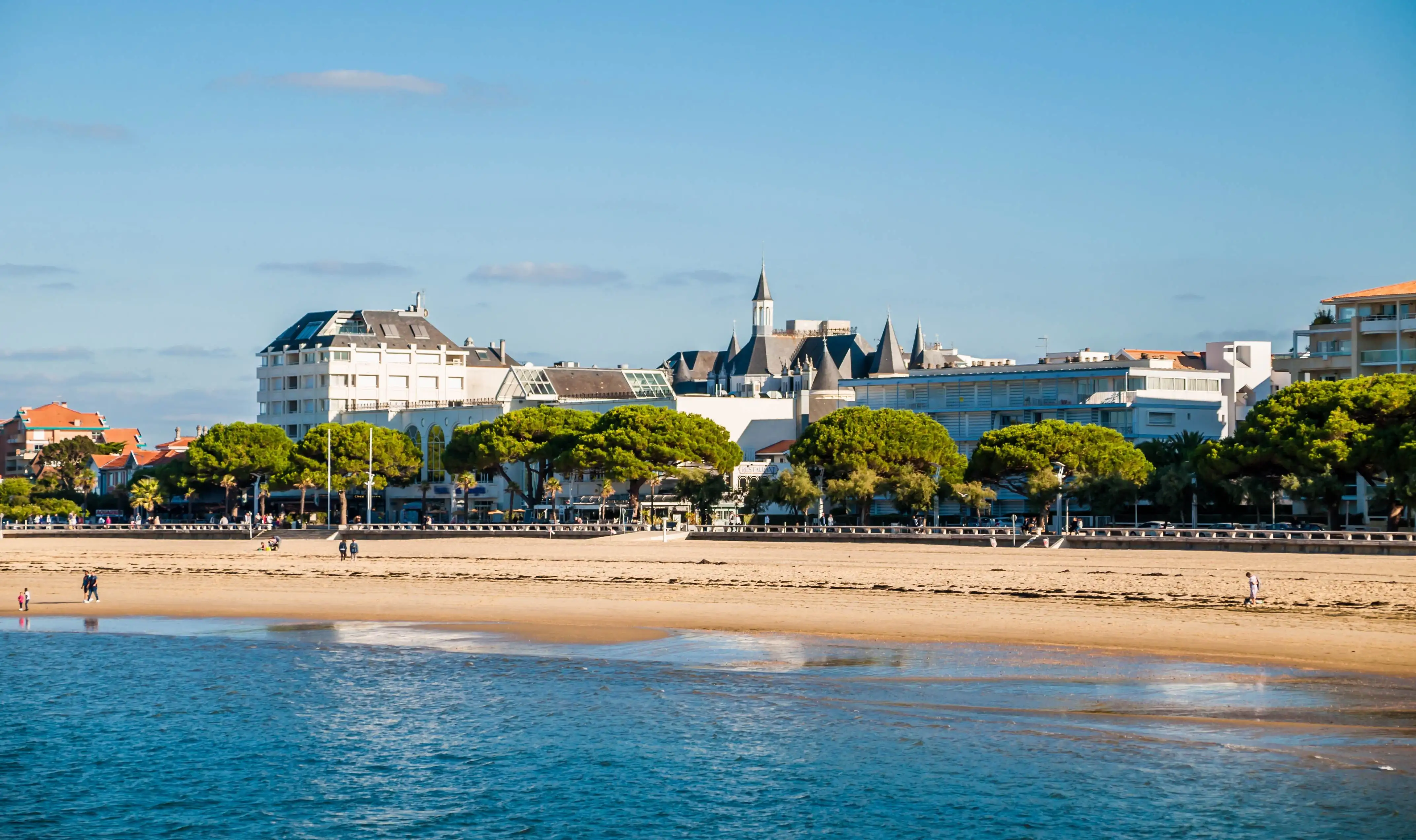 View of Arcachon, Gironde, from the sea on a clear blue day, with sandy shoreline and coastal buildings in the distance.