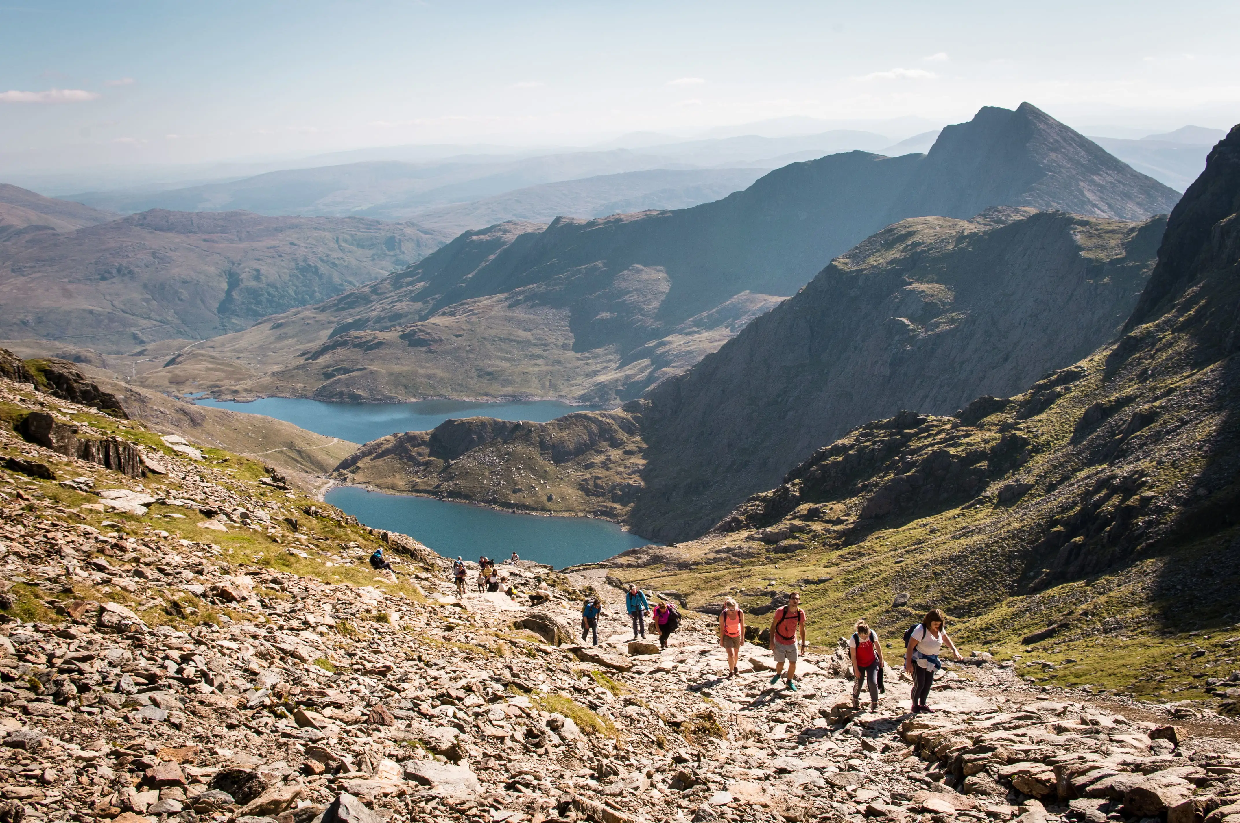 Bergwandelaars die over het rotsachtige pad van Mount Snowdon lopen, van bovenaf gezien met het indrukwekkende landschap van Wales op de achtergrond.