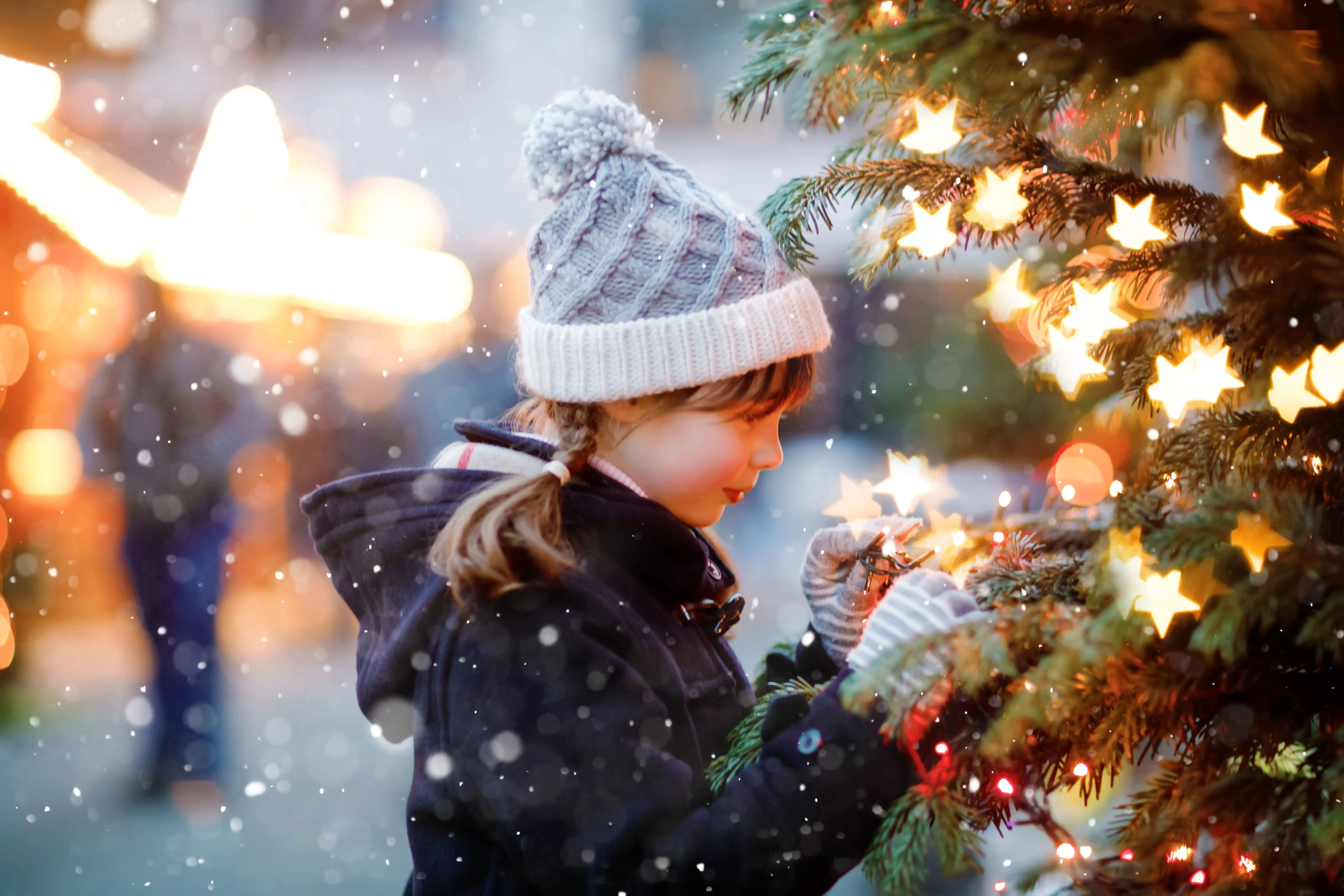 A young girl in a bobble hat looking enchanted by a Christmas tree, with snow in the air