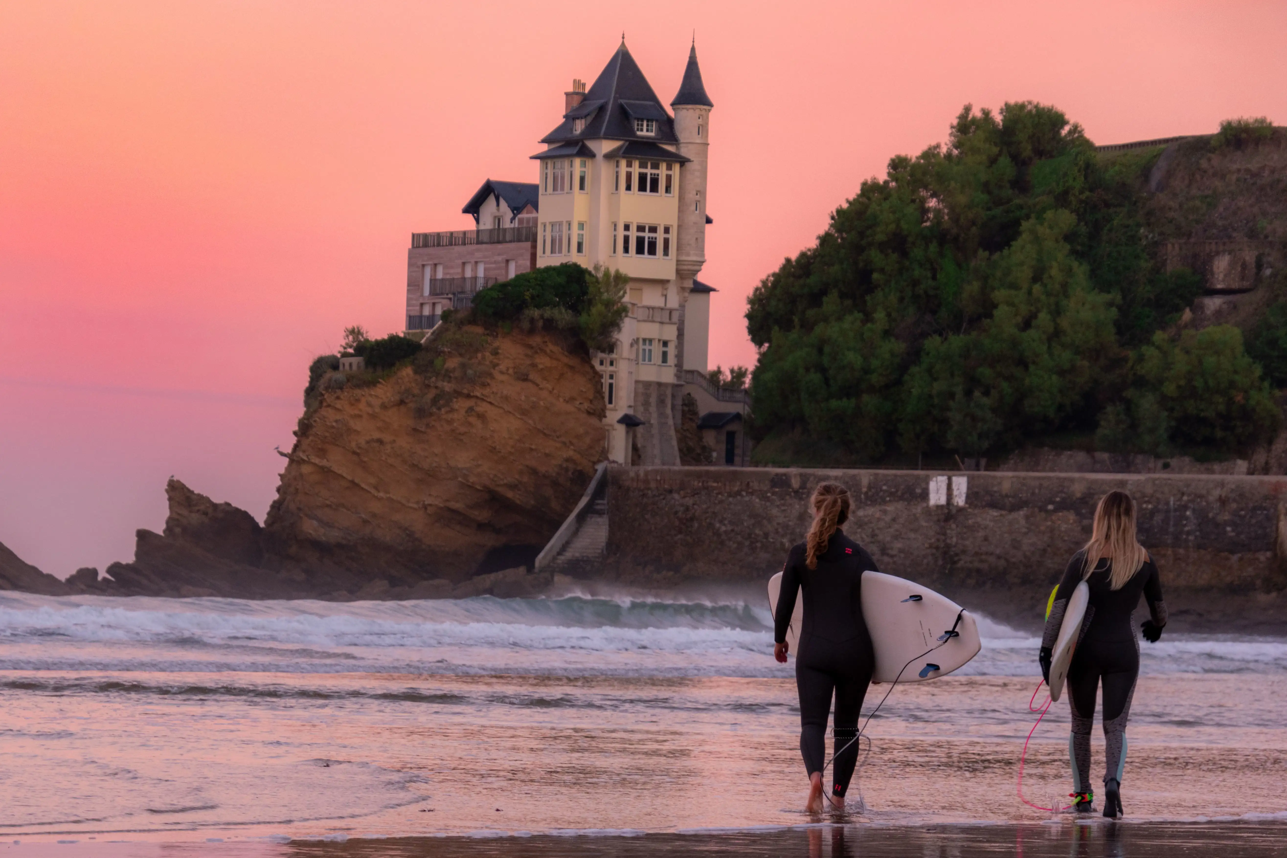 Surfers walking along the beach at sunset in Biarritz, carrying surfboards with Villa Belza visible in the background.