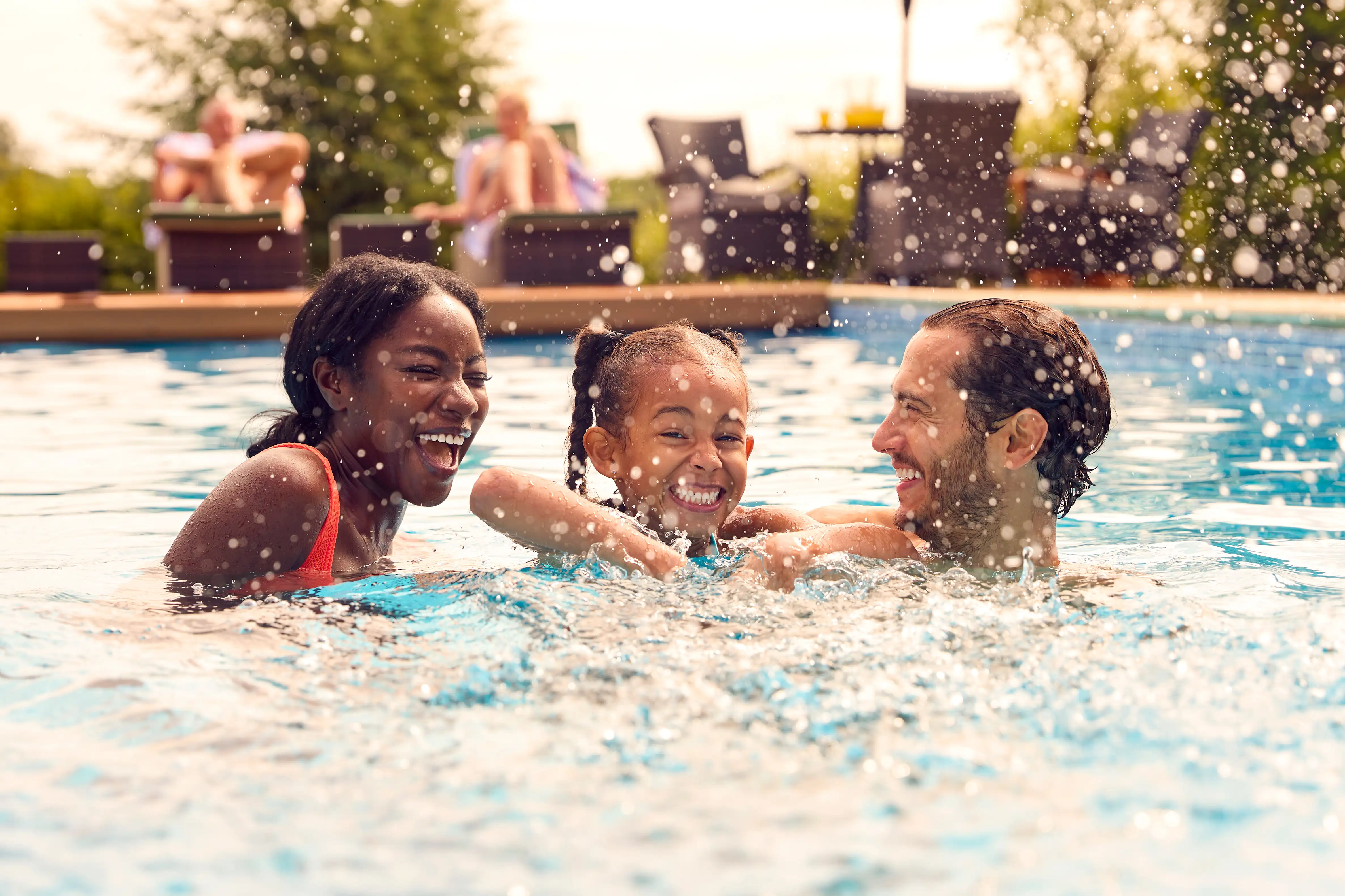 Family splashing and playing in a swimming pool with people relaxing in deckchairs in the background.