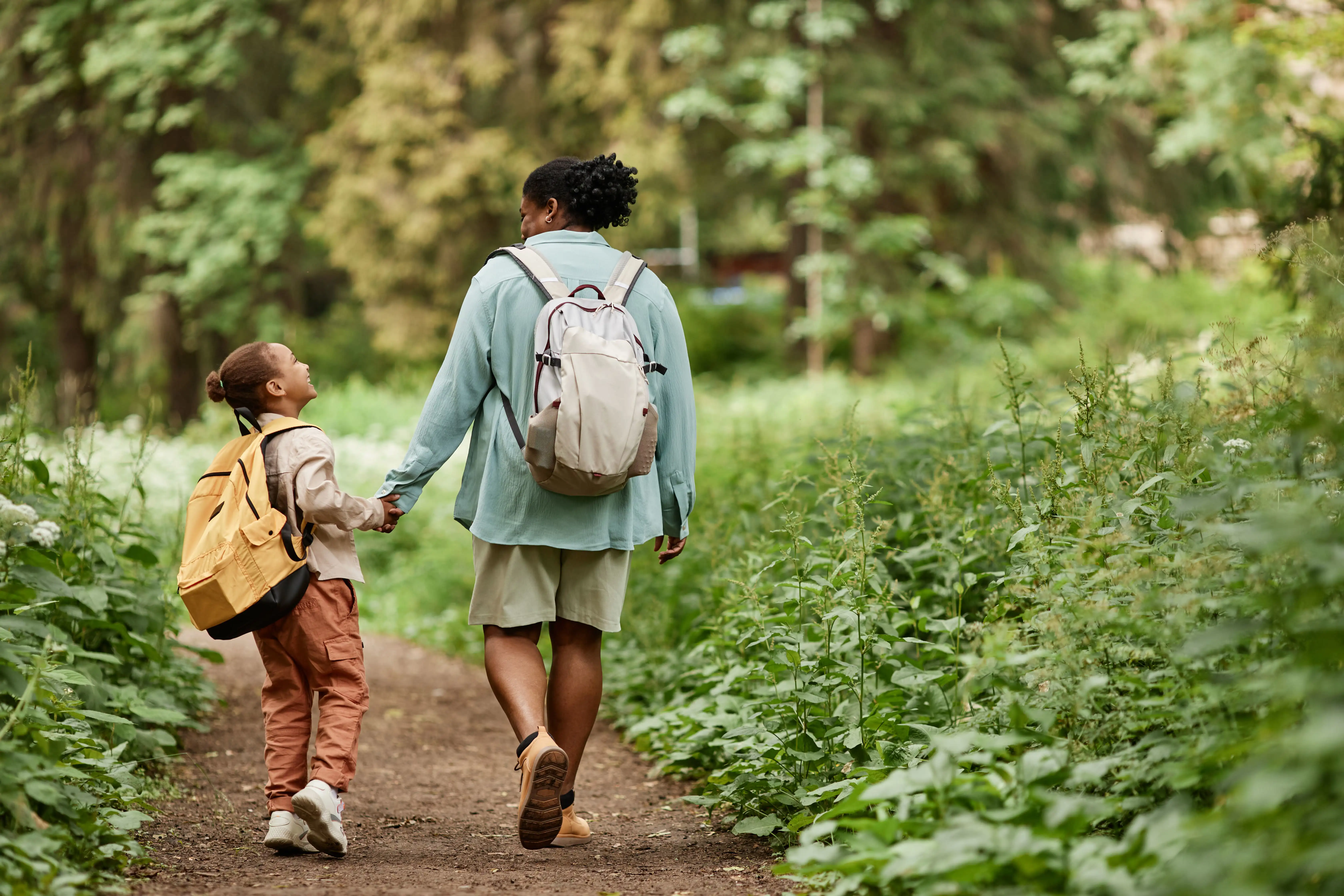 Mother and child with backpacks holding hands while walking through a forest trail.