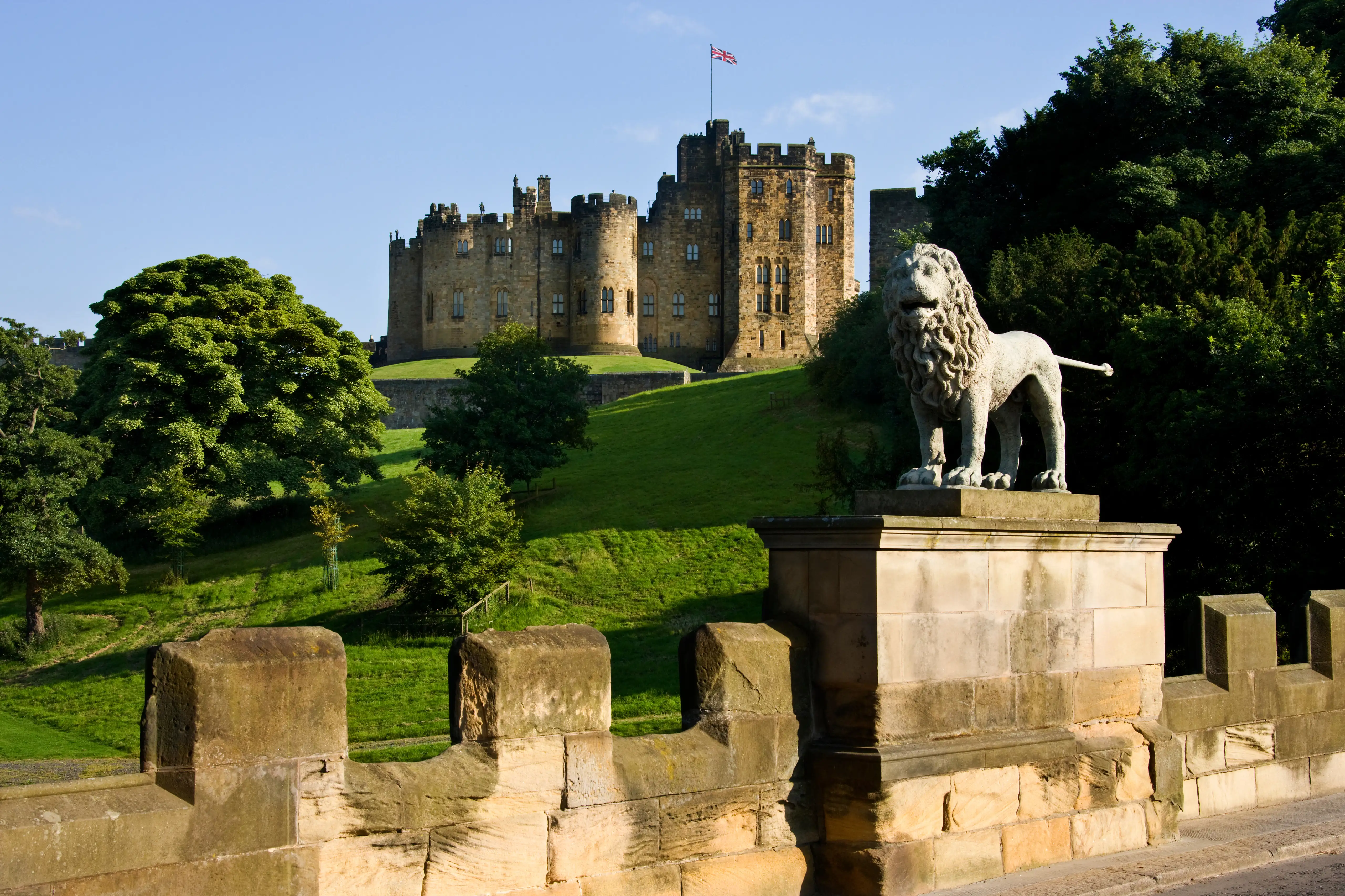 Alnwick Castle met zijn historische stenen muren, tegen een helderblauwe lucht, omgeven door groen gras en bomen.