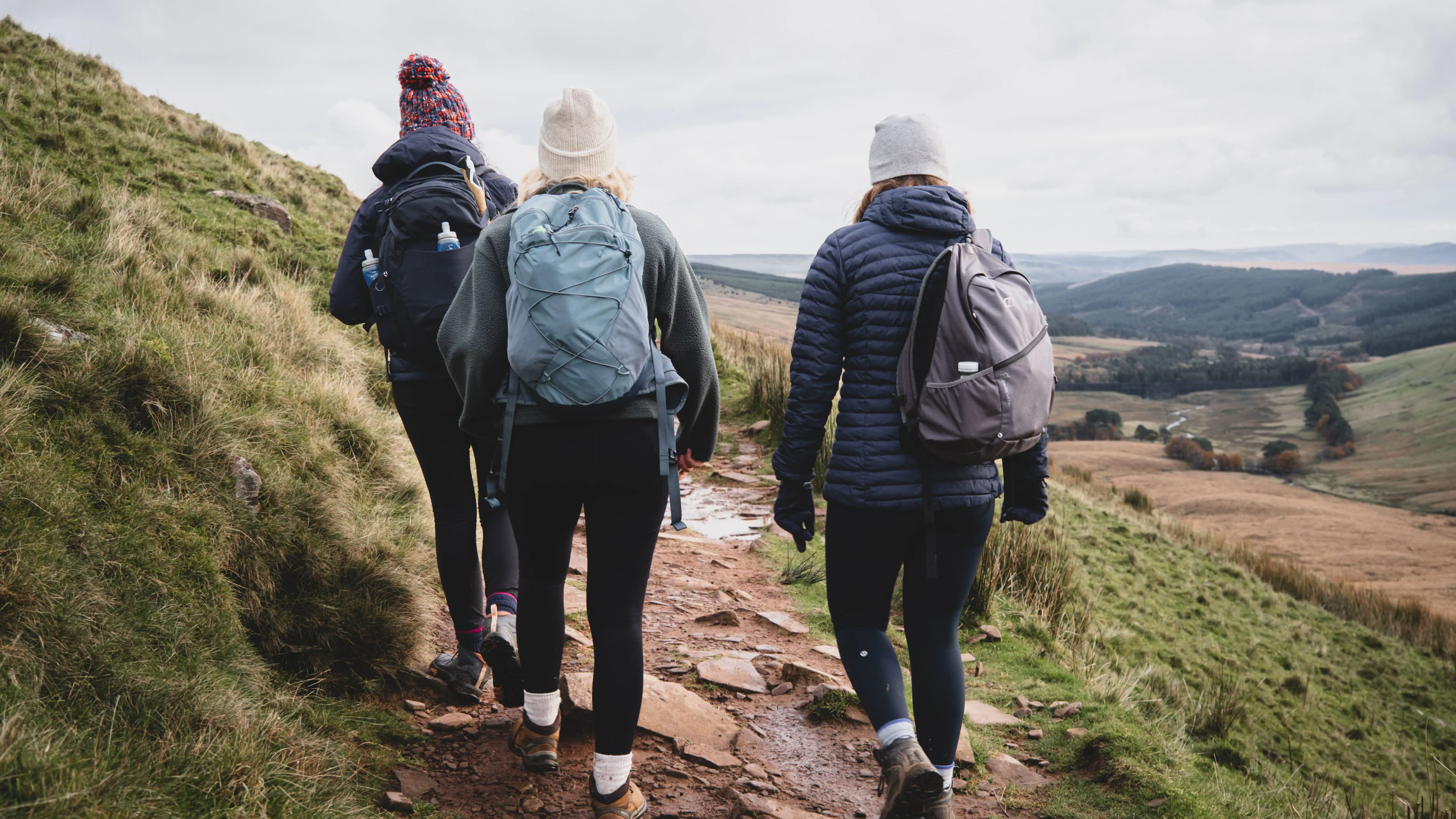 Drie warm aangeklede mensen lopen op een bewolkte dag bergopwaarts over een wandelpad, gezien vanaf de rugzijde.