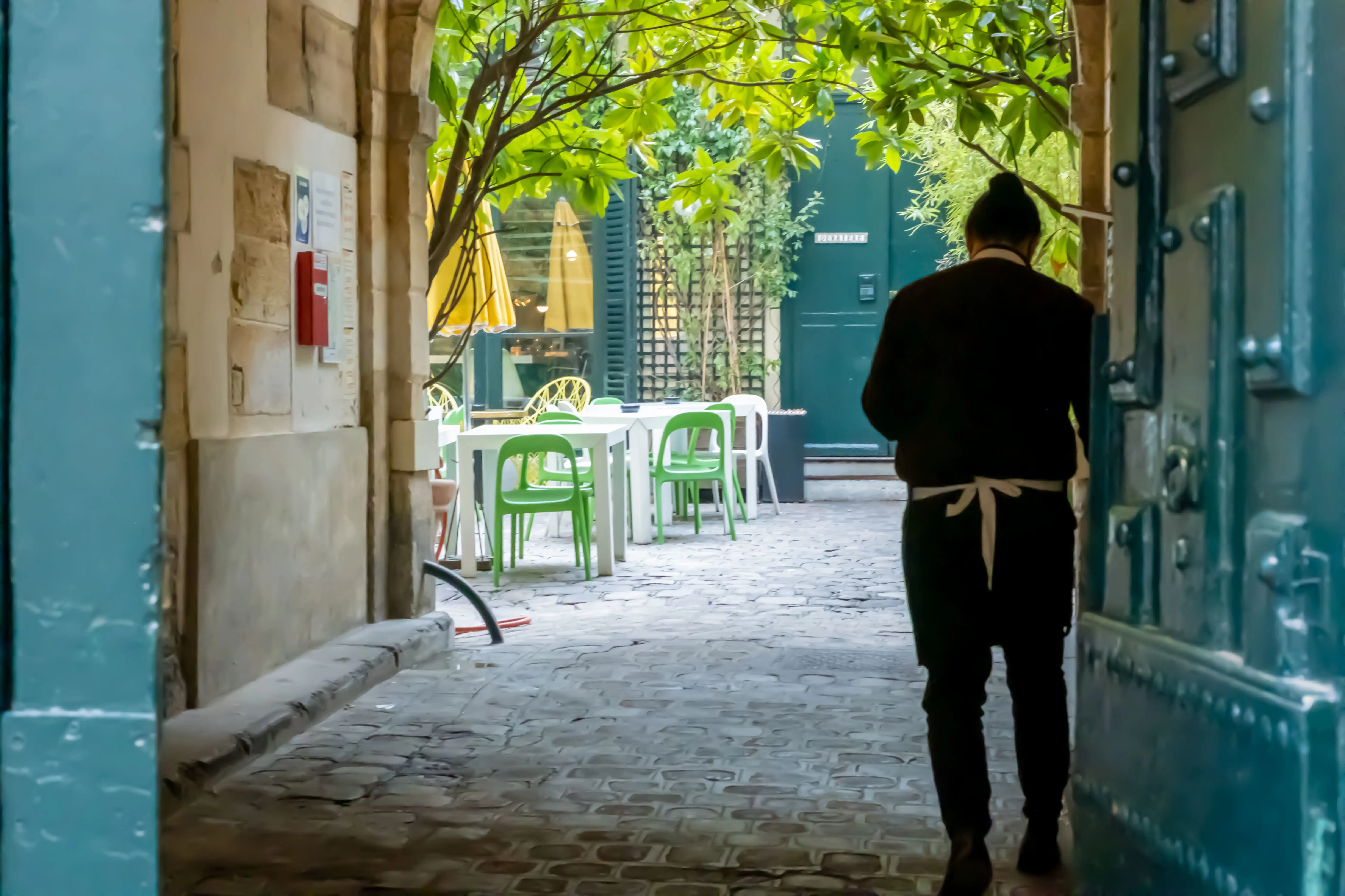 Restaurant tables in a hidden courtyard with a waiter with his back to a large doorway