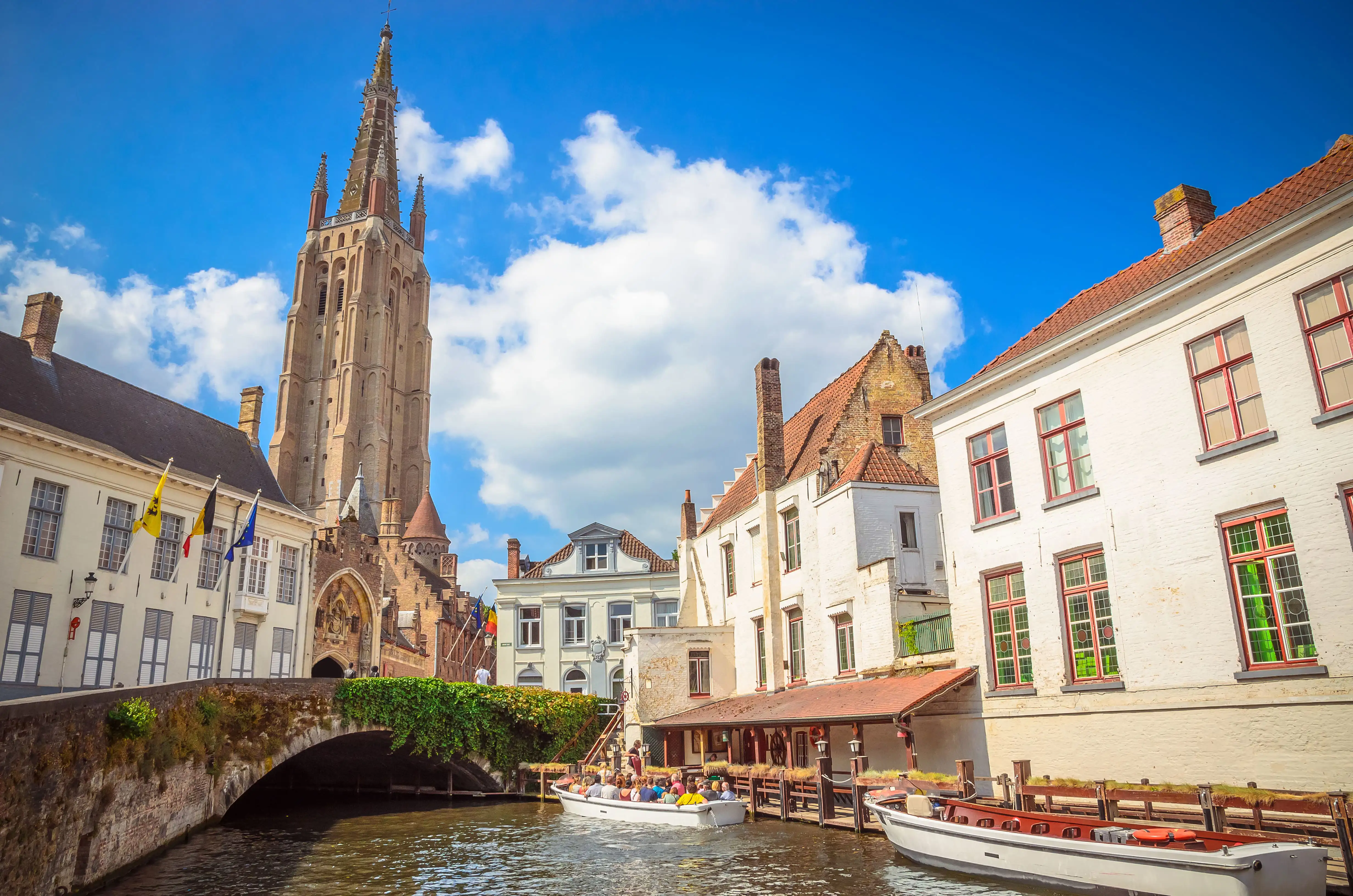 Church of Our Lady in Bruges seen from the canal, with boats in the water and attractive white buildings on either side of a bridge.