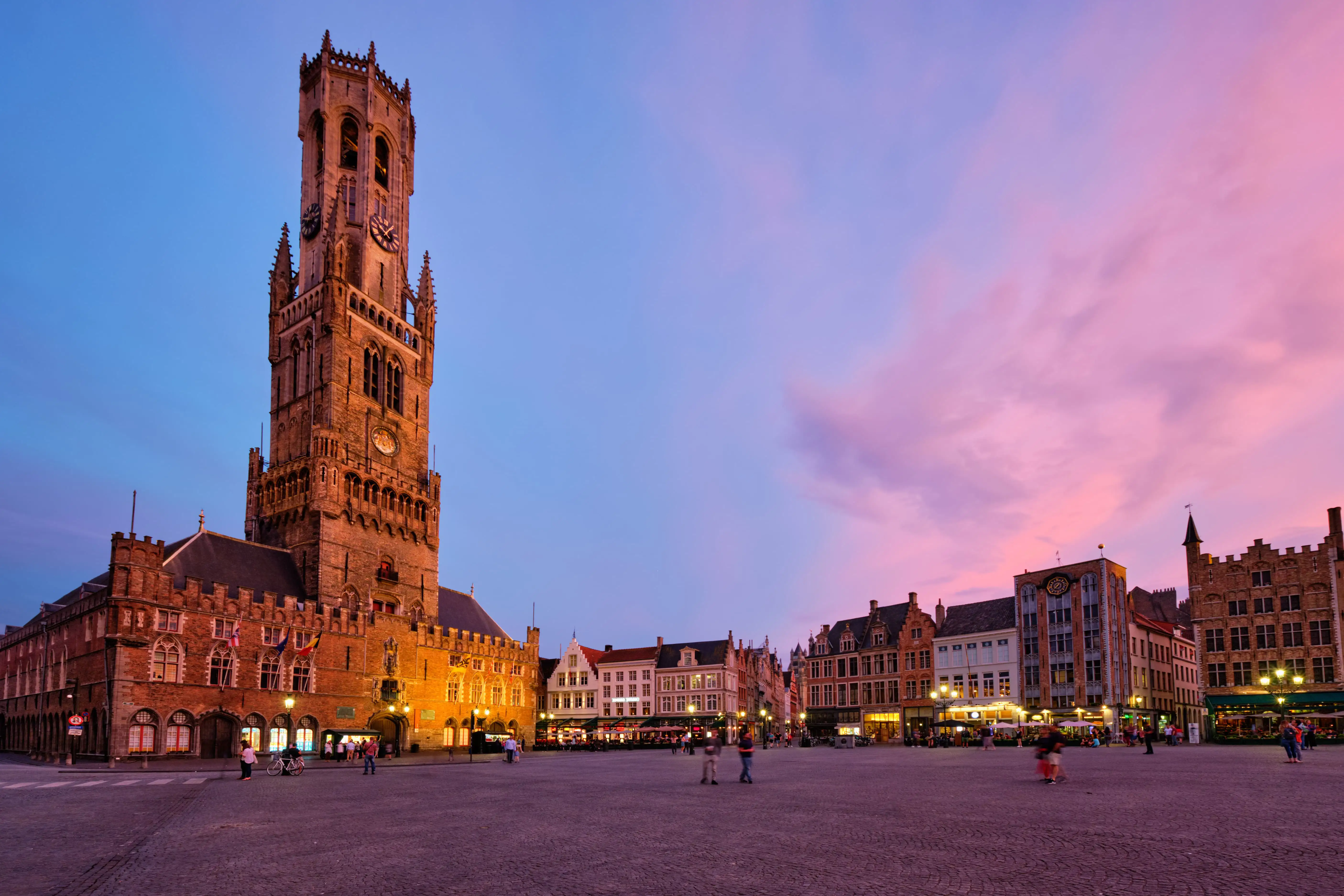 Belfry Tower in Bruges at sunset with pink and blue sky and warm city lights.