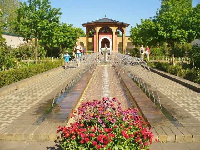 A brick path leads to a yellow and red pavilion. Along the centre of the path is a water feature with individual streams of water arching up and toward the centre. Trees line the perimeter and families are walking by. Pretty red flowers are in the forefront of the image.