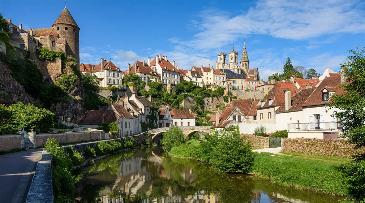 A panoramic view of the medieval town of Semur-en-Auxois, with its fortified walls and ancient stone towers rising above a river and bridge.