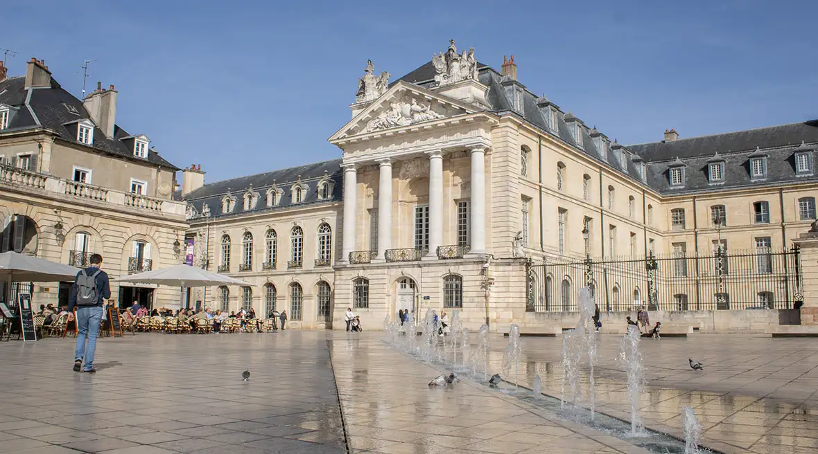 The Palace of the Dukes of Burgundy, a grand historic building, towers over the open Place de la Libération, with fountains and outdoor cafe seating in the foreground.