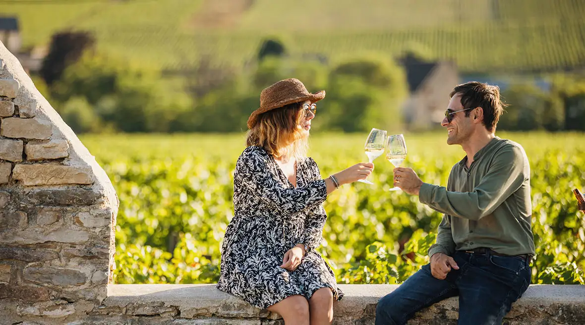 A man and a woman toast with glasses of white wine while sitting on a low stone wall, with rows of grapevines stretching out behind them under a bright sky.