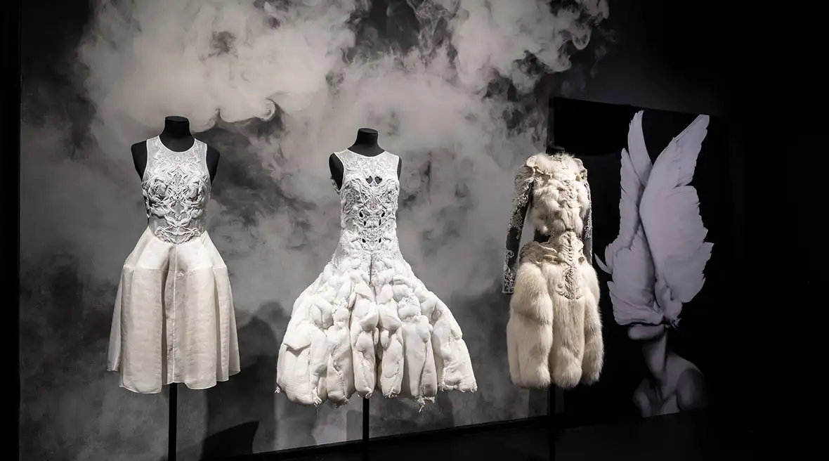 A display of three all-white dresses from the Yiqing Yin "Air and Dreams" exhibit. The garments are set against a background of white smoke and a black and white image of a person with large white wings