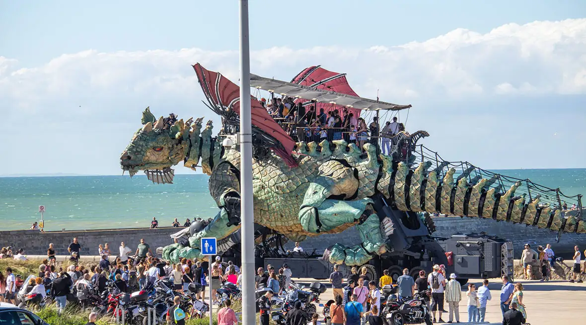 A massive mechanical dragon with people riding on its back stands near a crowd on a promenade by the beach in Calais