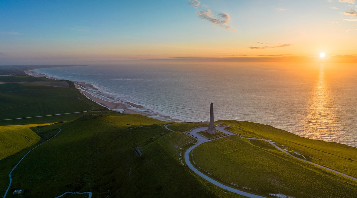 clifftop overlooking a vast ocean with a vibrant orange sunset on the horizon and a curving coastline stretching into the distance