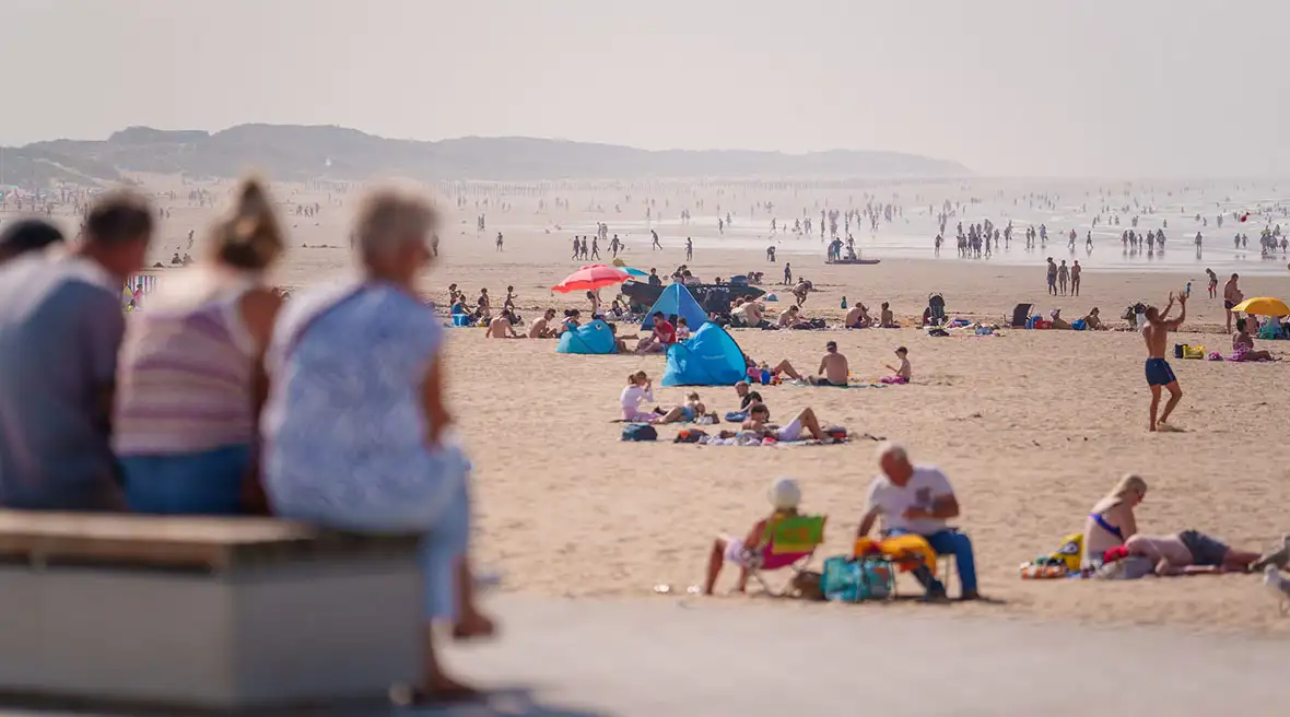 A crowded beach on a sunny day with many people relaxing on the sand. In the foreground, three people are out of focus, sitting on a bench