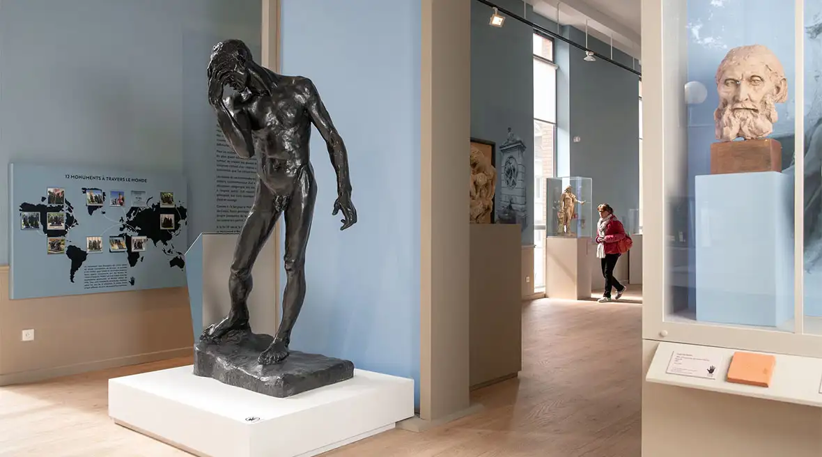 A statue of a distressed, muscular man by Auguste Rodin is displayed in the foreground of a museum gallery