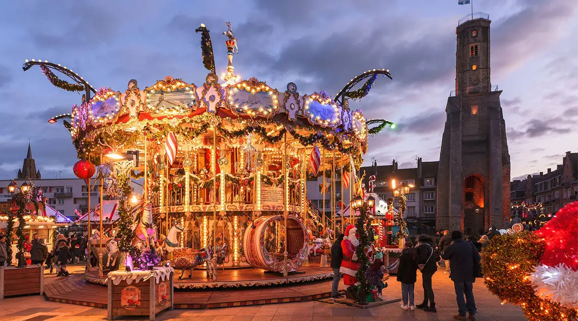 A lit carousel decorated for Christmas is the centerpiece of a festive market square at dusk. In the background, a historic tower stands tall