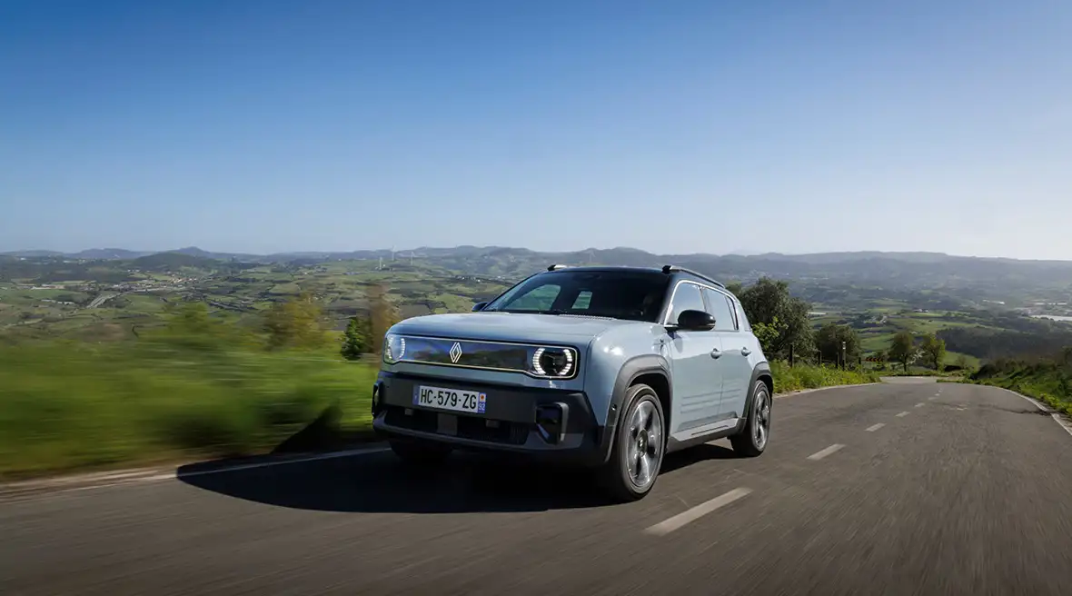 A light blue Renault 4 electric SUV driving on a winding countryside road with scenic hills and a clear blue sky in the background