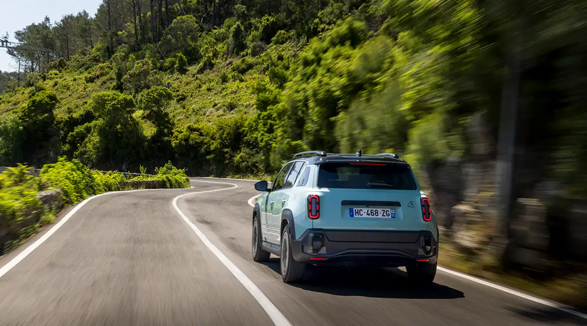Rear view of a light blue Renault 4 electric SUV driving on a curvy road through a lush, forested landscape