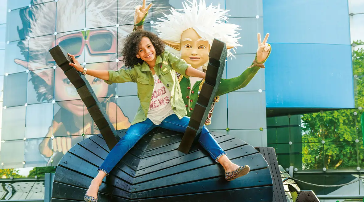 A happy young girl sits in front of a theme park statue of an animated film character.