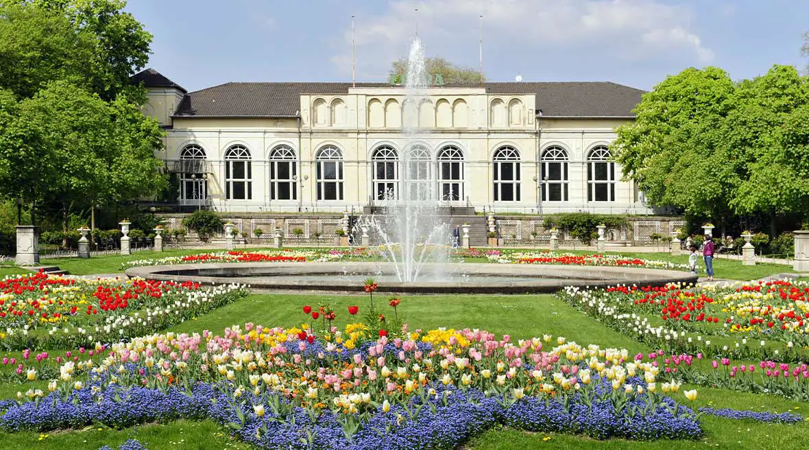 White building with green trees either side of it with green lawns and flower beds and fountain.