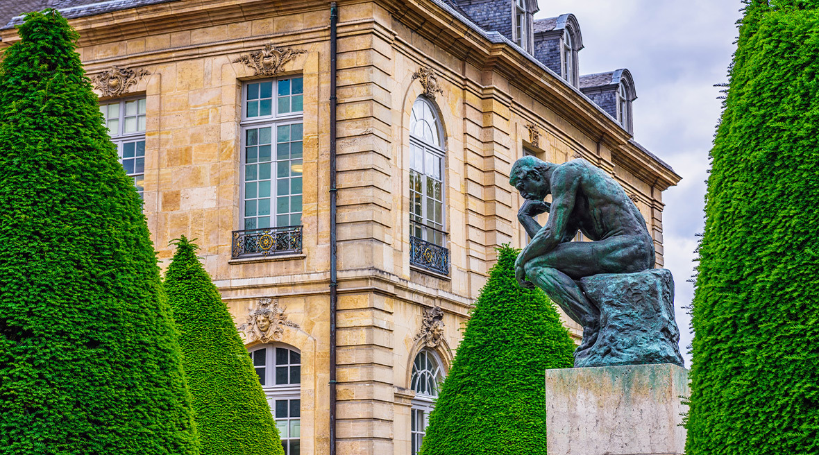 Statue of Rodin's 'The Thinker' in an ornamental garden in Paris with a grand building behind.