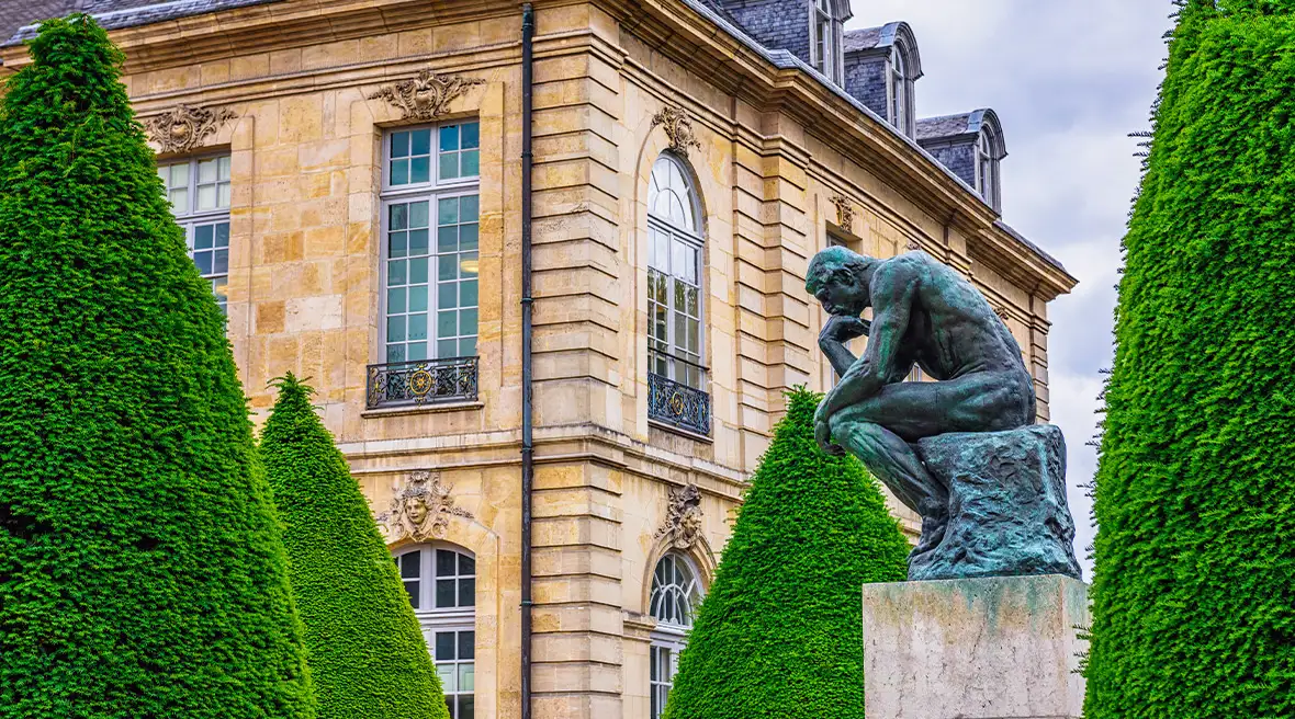 Statue of Rodin's 'The Thinker' in an ornamental garden in Paris with a grand building behind.