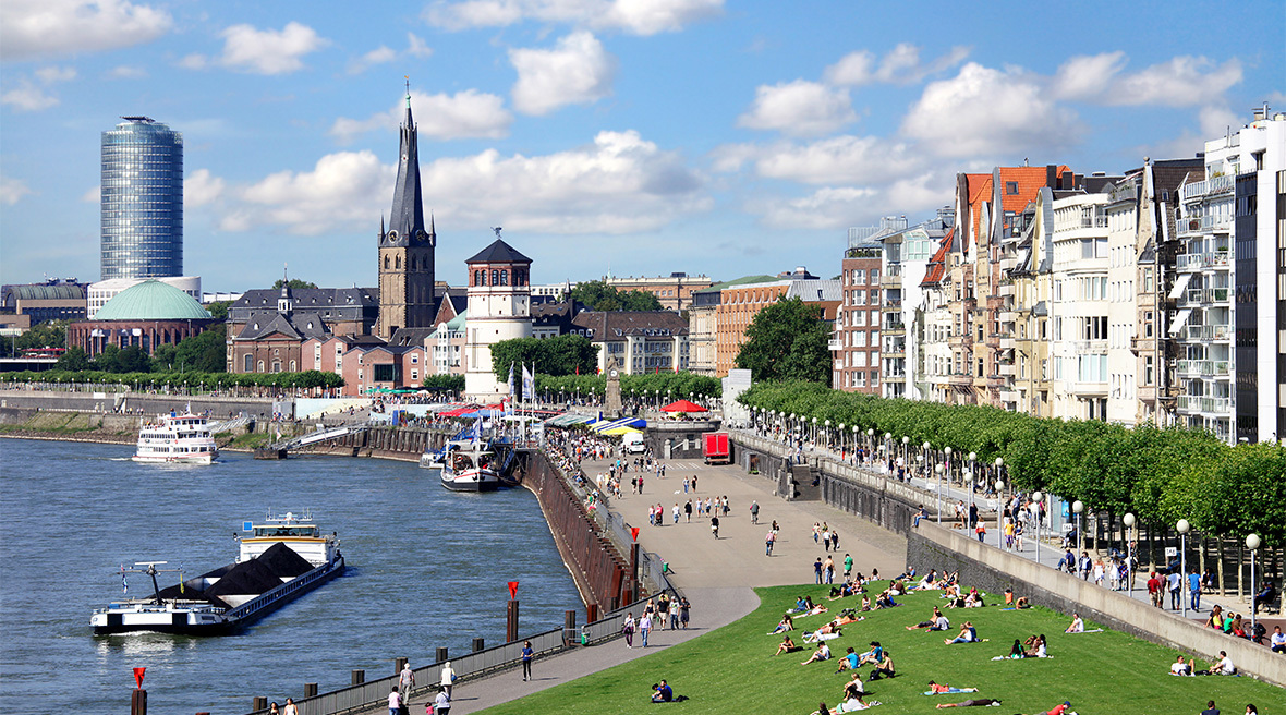 A wide walkway beside a river in a city, with green space in the foreground