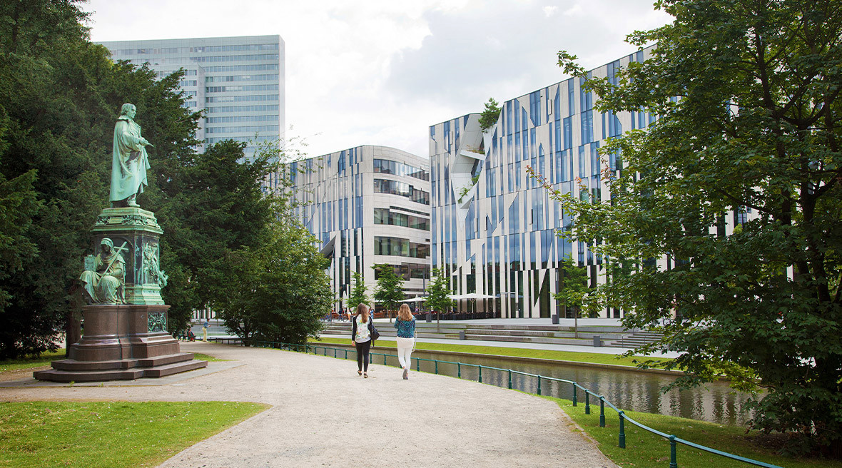 Two people walking in a city park by a green statue towards a modern building