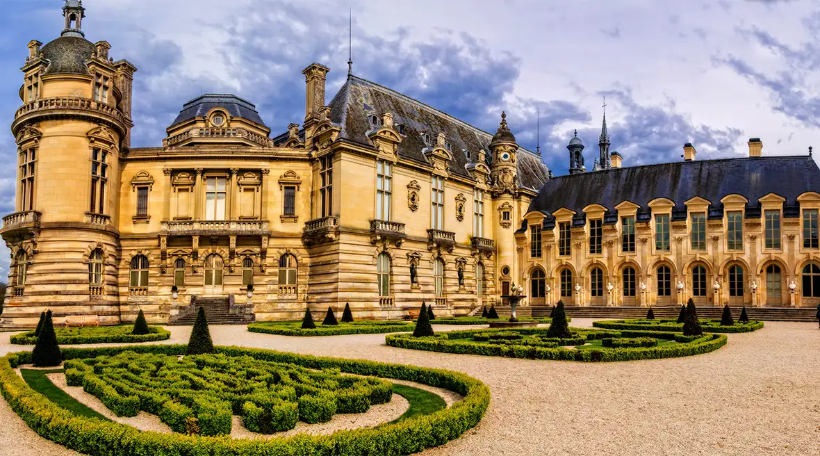 An elegantly decorated château on three floors with tall windows stands behind a large gravel drive with ornamental borders