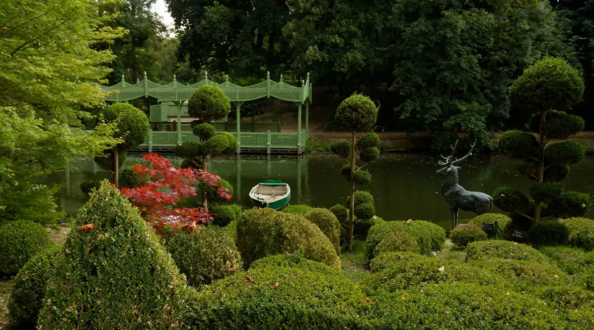 Manicured hedges in foreground, with an ornamental lake behind and a statue of a deer overlooking the water and one boat in the lake