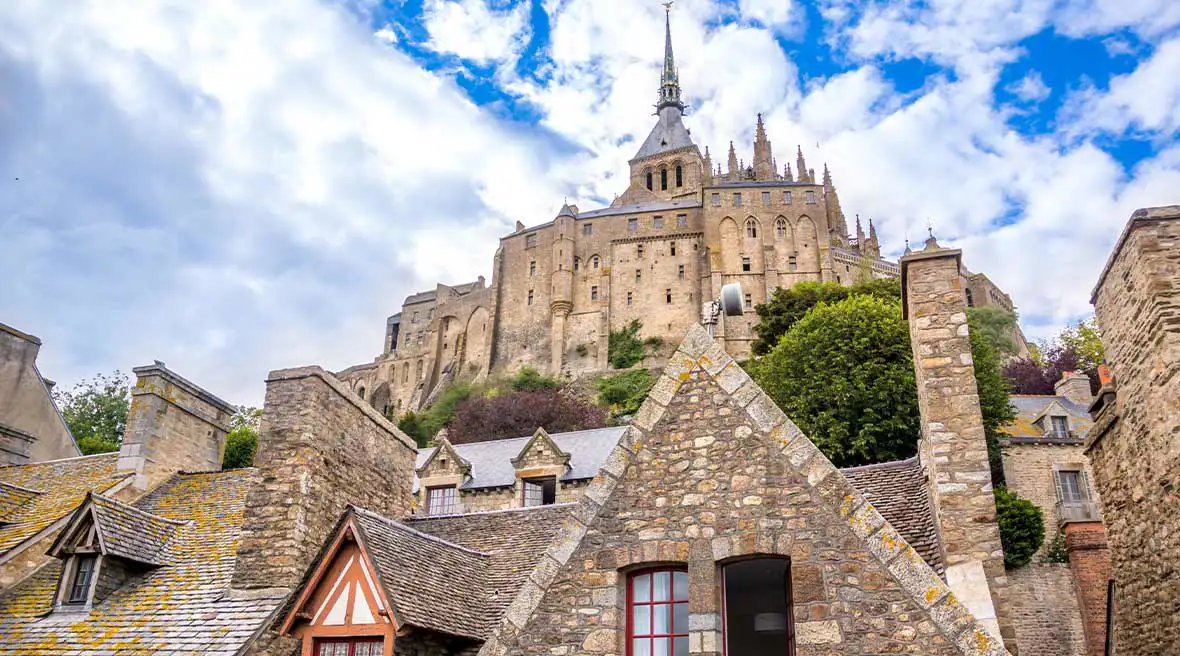 Yellow and grey stone building with one open window. Behind the building is a green tree and the Abbey on the hill beneath a blue sky with white clouds