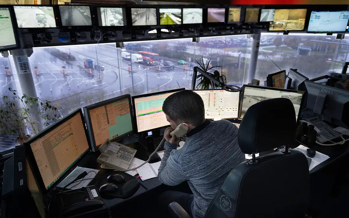 Man in blue shirt operating a computer with 5 screens in a control room full of screens keyboards and controls