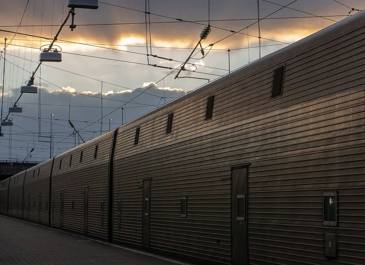 The outside of a row of train carriages all grey metal with small windows and doors dotted along pulled in at station