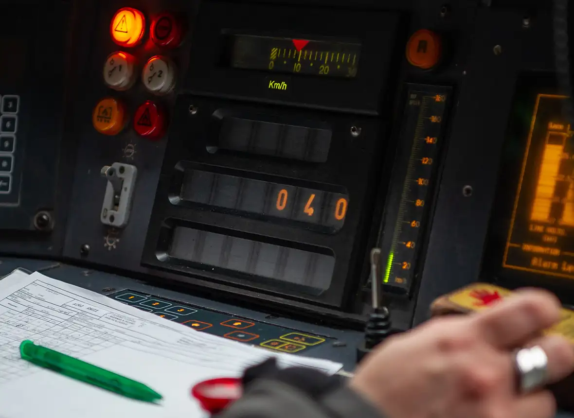Close up of a driver’s hand in a train with a green pen and paper in front of them with the screen and controls that display the train’s speed and signal controls